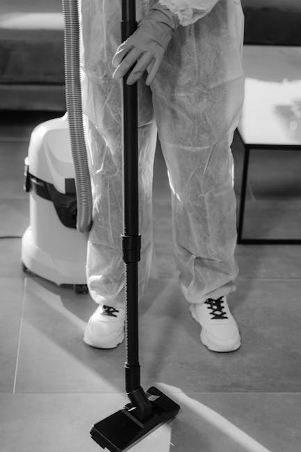 A person in protective clothing, including gloves and a disposable suit, is using a vacuum cleaner with a long flexible hose and a wide floor attachment to perform surface cleaning in an indoor space.