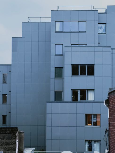 Exterior view of a modern multi-storey building with a grey facade composed of large, smooth panels. Several windows, some reflecting the sky, are visible, with a mixture of open and closed blinds. The building's clean and sleek appearance suggests recent maintenance or cleaning, possibly by professionals such as those at Cleaners SW3. The image emphasizes the building's structure and design details, with natural lighting highlighting the surface cleanliness, indicative of surface cleaning and ongoing building upkeep relevant to residential or commercial property maintenance.