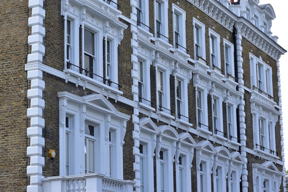 Photograph of the exterior façade of a Victorian-style residential building on Kings Road Chelsea, showcasing decorative white window frames, ornate cornices, and balustrades accentuating large sash windows on a brick wall. The building features multiple floors with tall, narrow windows, some with black iron railings, and is lit by natural daylight. The surface materials include brown brickwork and white decorative woodwork that appears clean and well-maintained, reflecting professional exterior cleaning, surface polishing, and preservation services provided by Cleaners SW3. The image emphasizes the building's architectural detail and tidy appearance, suitable for illustrating residential cleaning and maintenance in the SW3 district.