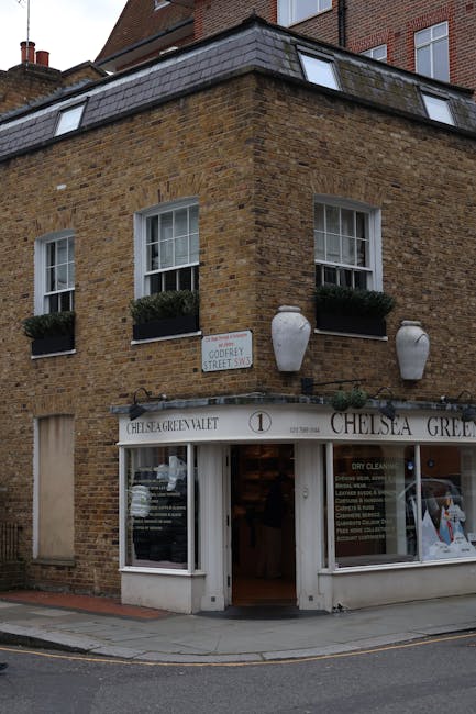 Exterior view of a corner building in Chelsea with a traditional brick façade, featuring three large white planters with green foliage attached to the upper windows. The ground floor houses a cleaning service shop, with a white sign reading 'CHELSEA GREEN VALET' and large glass windows displaying cleaning service information. The entrance is open, revealing a clean, well-lit interior. The street is paved and appears tidy, with a street sign indicating Gough Street. Natural daylight illuminates the scene, highlighting the building's neat appearance. As part of the cleaning services offered by Cleaners SW3, surface cleaning, deep cleaning, and sanitisation of residential and commercial spaces in Chelsea are expertly managed, ensuring high standards of hygiene and shine.
