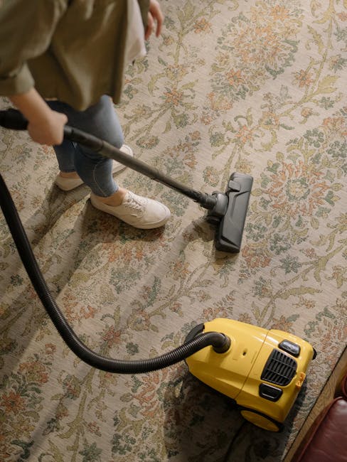 A person operates a yellow vacuum cleaner with a black hose attachment on a traditional patterned area rug, situated in a well-lit room with wooden flooring and furniture. The individual is dressed in casual attire, wearing beige sneakers and jeans, engaged in surface cleaning. The rug features a floral motif with shades of cream, green, and orange, and appears clean and well-maintained. The scene emphasizes professional upholstery cleaning processes, aligning with services offered by Cleaners SW3 for deep cleaning and stain removal in Sloane Square Chelsea. The image showcases the use of a portable vacuum device to enhance hygiene and cleanliness for domestic or commercial spaces.