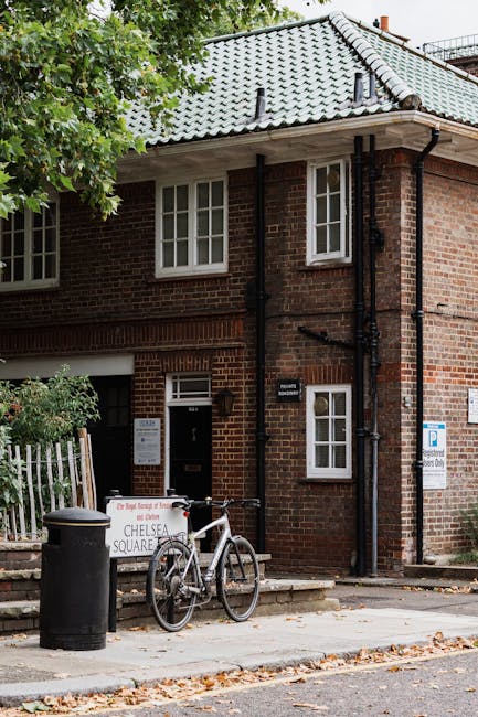 Exterior view of a brick residential building with a tiled, slate-gray roof, situated on a tree-lined street in Chelsea. The building features white-framed windows and a black door with a small window. In front, there is a bicycle resting against a railing, a black trash bin, and a sign indicating parking regulations. The scene is well-lit with natural daylight, and the street has fallen leaves scattered on the pavement. The image represents a typical Chelsea flat's outside environment, highlighting cleanliness and maintenance, relevant to domestic and surface cleaning services offered by Cleaners SW3 as part of end of tenancy cleaning checklists for Chelsea flats.